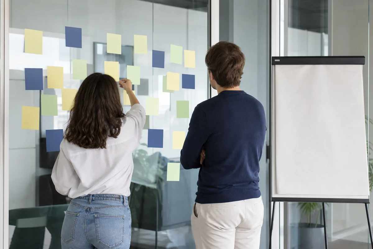Deux collègues examinent des post-it colorés sur une vitre lors d&rsquo;un atelier de cadrage