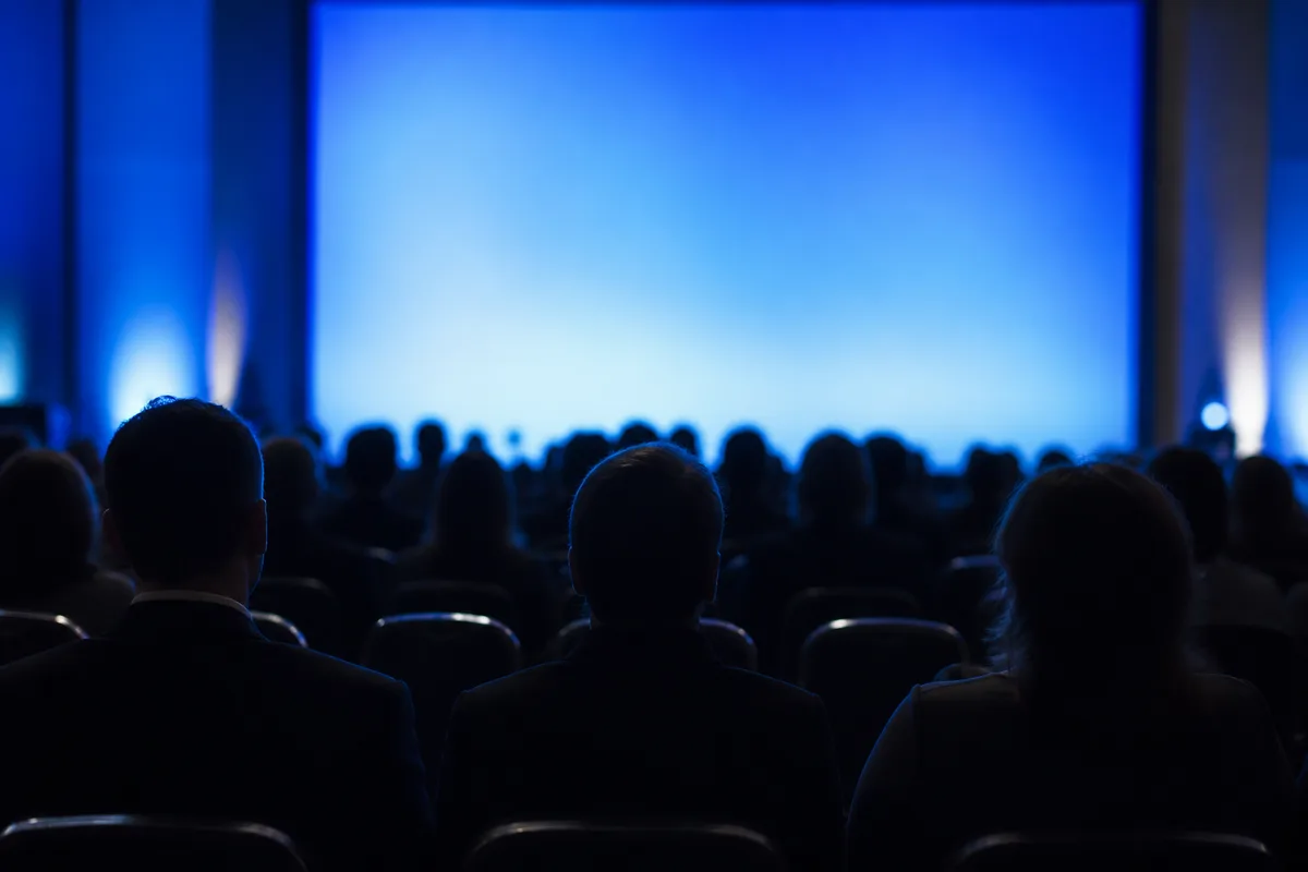 Salle de conférence vue de dos pendant une présentation, sièges occupés en silhouette, écran lumineux en arrière-plan flou